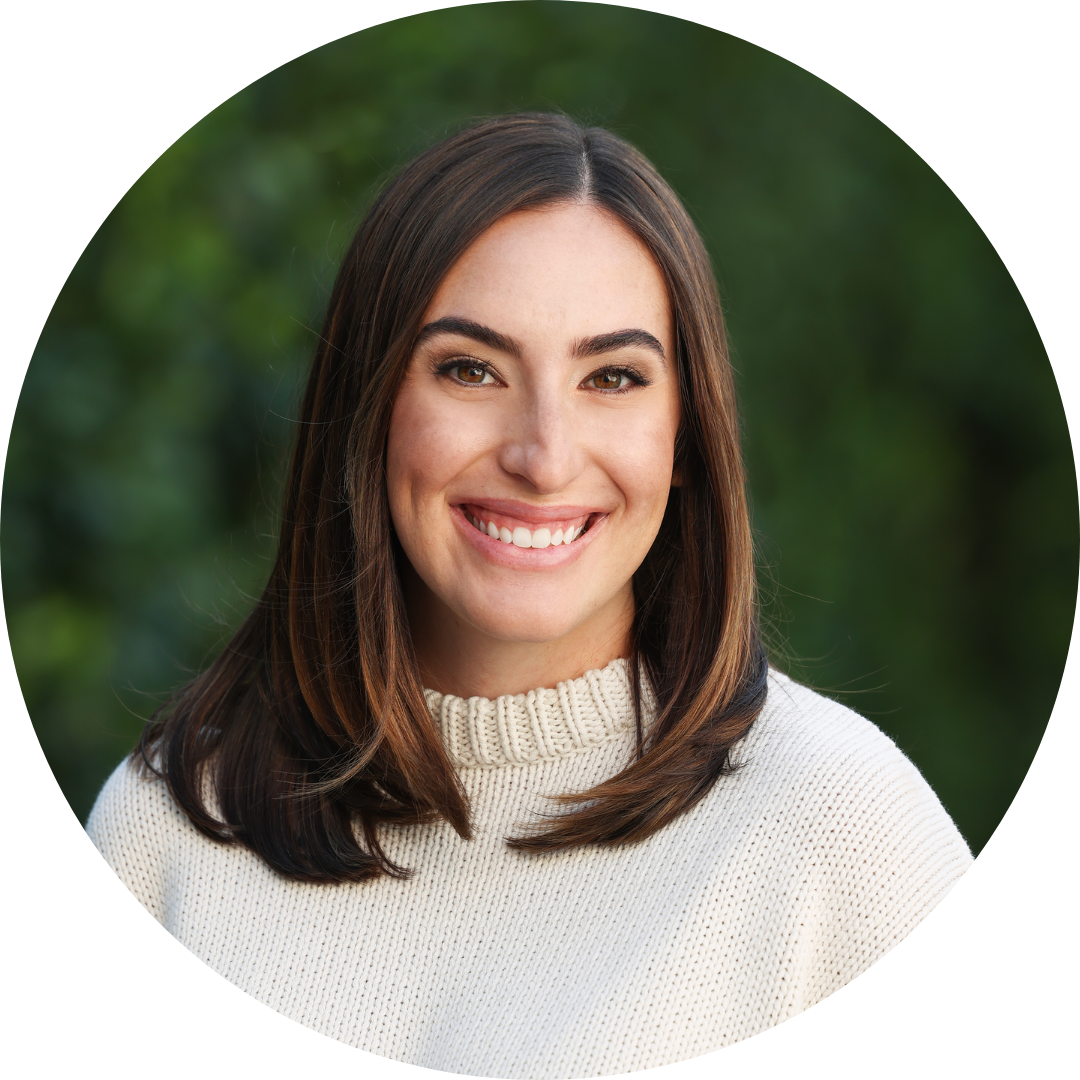 A woman with straight brown hair, wearing a cream-colored sweater, smiles at the camera in front of a blurred green background.