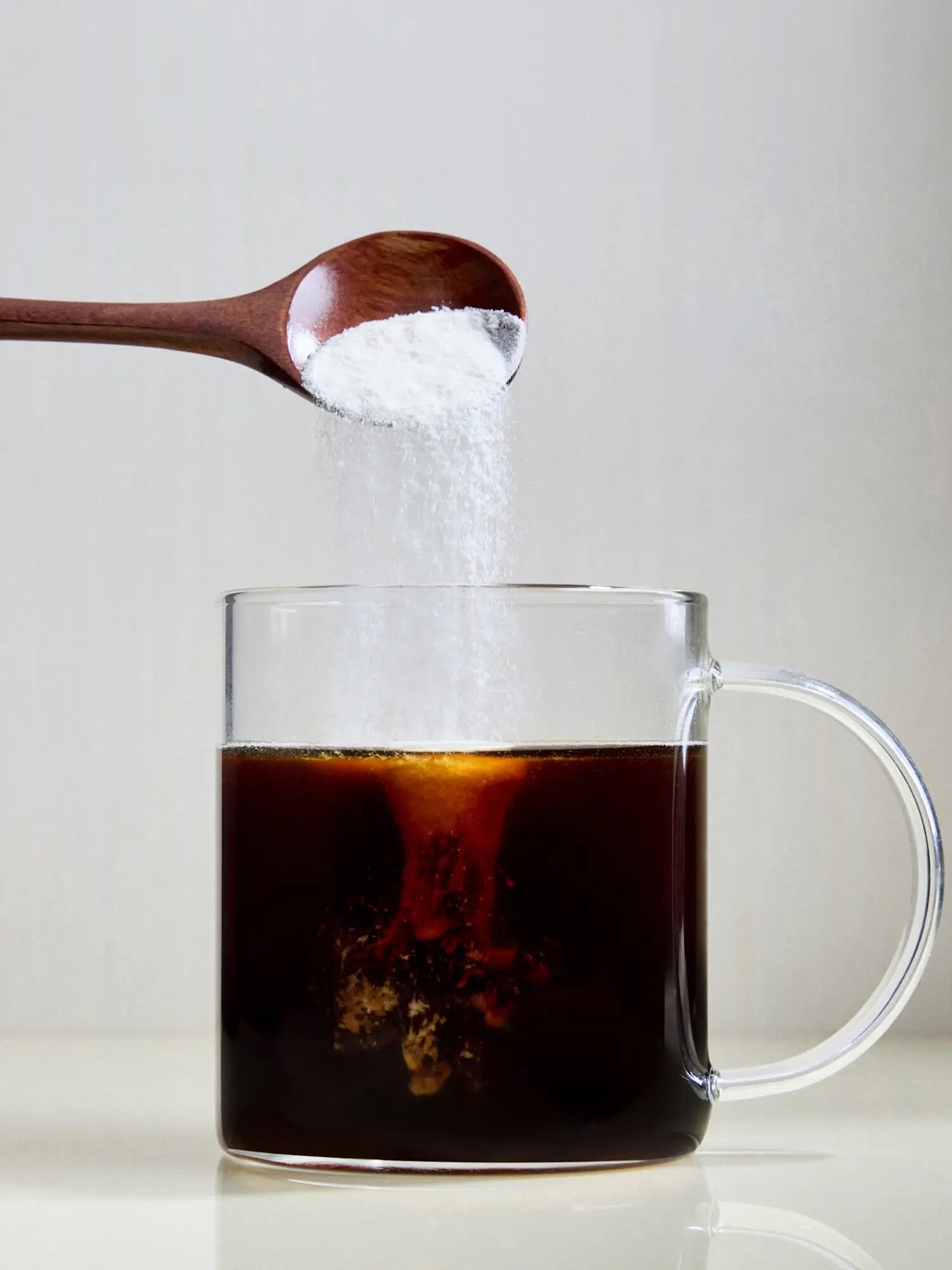 A spoon is pouring white sugar into a clear glass mug filled with black coffee against a plain background.