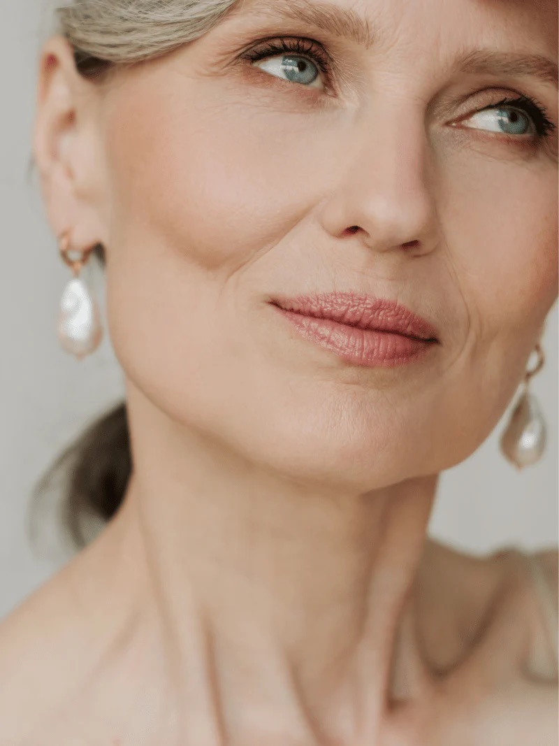 Close-up of a middle-aged woman with fair skin, light eyes, and gray hair wearing pearl drop earrings, gazing slightly to the side with a neutral expression.