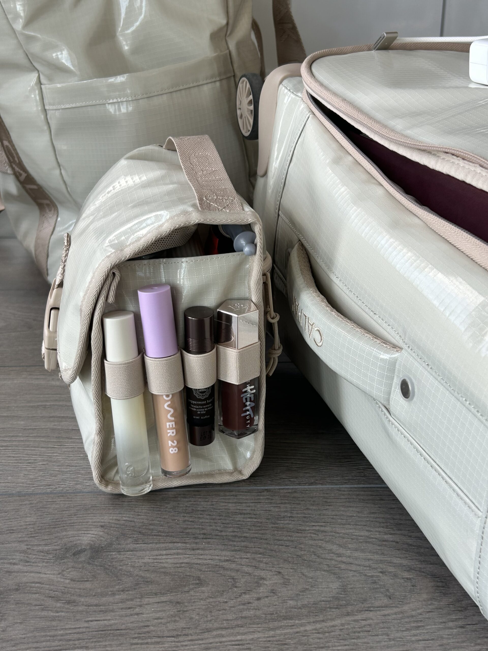 A small beige toiletry bag with assorted cosmetic products stands open on a wooden floor next to two matching beige travel bags.