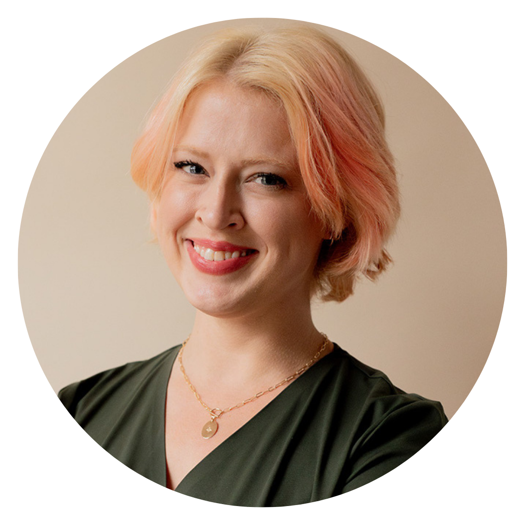 A person with short, light blonde hair with pink highlights, wearing a dark top and a gold necklace, smiling at the camera against a neutral background.
