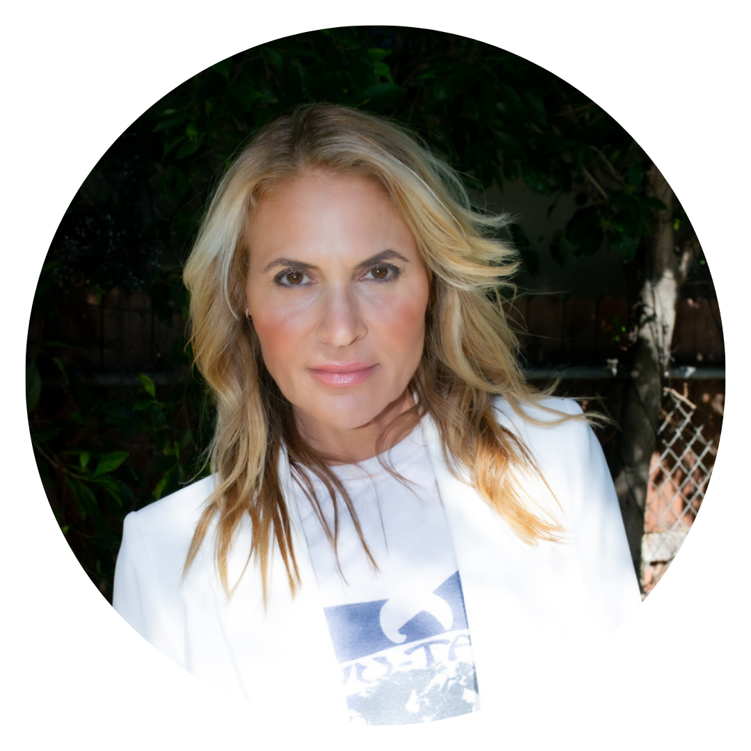Woman with long blonde hair wearing a white blazer and graphic t-shirt, standing outdoors in front of greenery and a chain link fence.