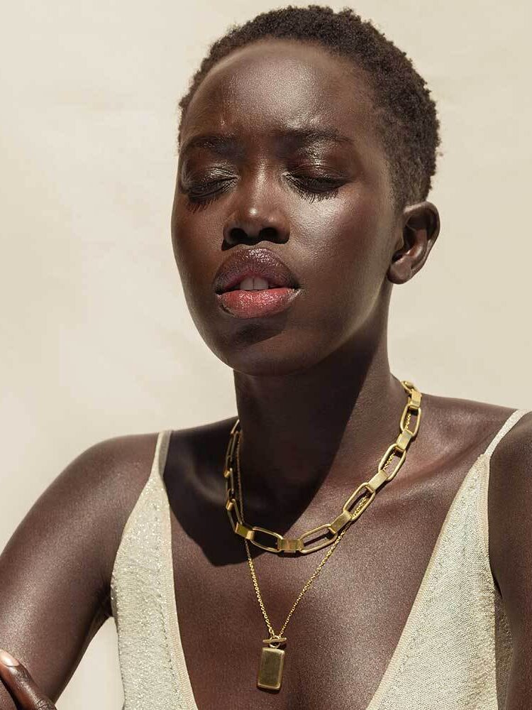 A woman with short hair and dark skin wears a white sleeveless top and a chunky gold chain necklace, posing with her eyes closed against a plain, light background.