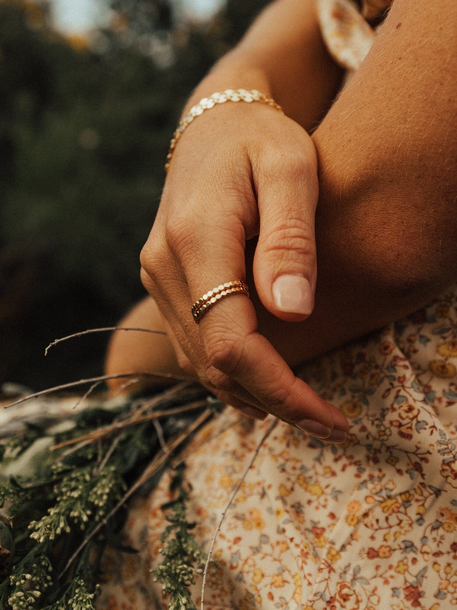 A close-up of a person's hand wearing gold jewelry, holding a small bouquet of wildflowers while wearing a floral-patterned dress.