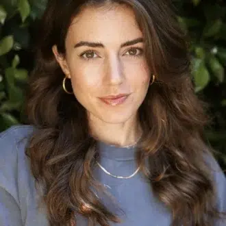 Woman with long wavy brown hair wearing a blue top, gold hoop earrings, and a gold necklace, standing in front of leafy green foliage.