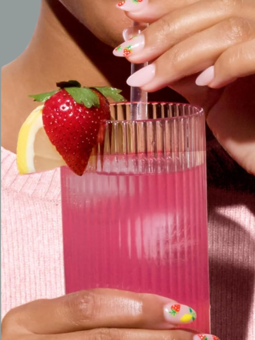 A person sips a pink beverage through a straw from a ribbed glass, garnished with a strawberry and lemon slice, with strawberry-themed nail art visible.