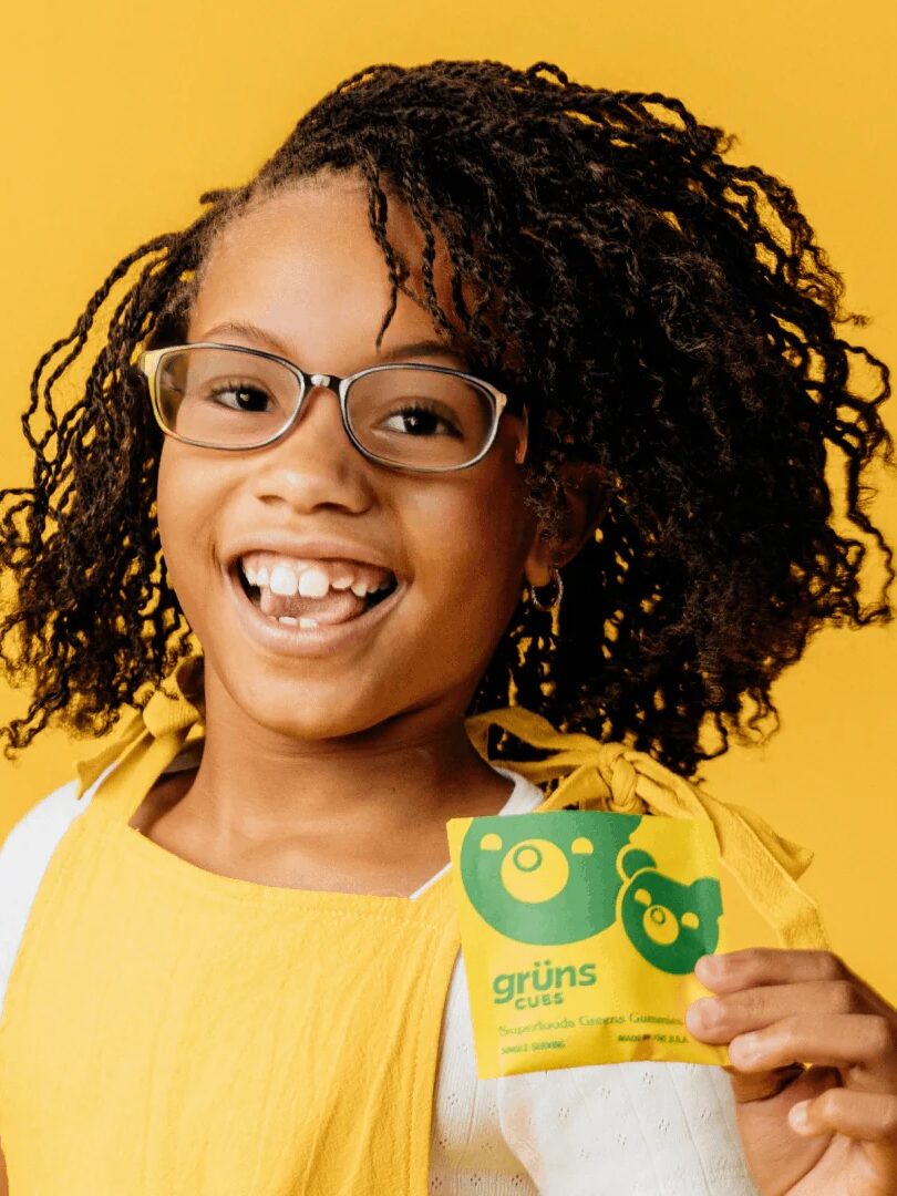 Smiling child with glasses and curly hair holds a green and yellow "grüns cues" packet against a solid yellow background.