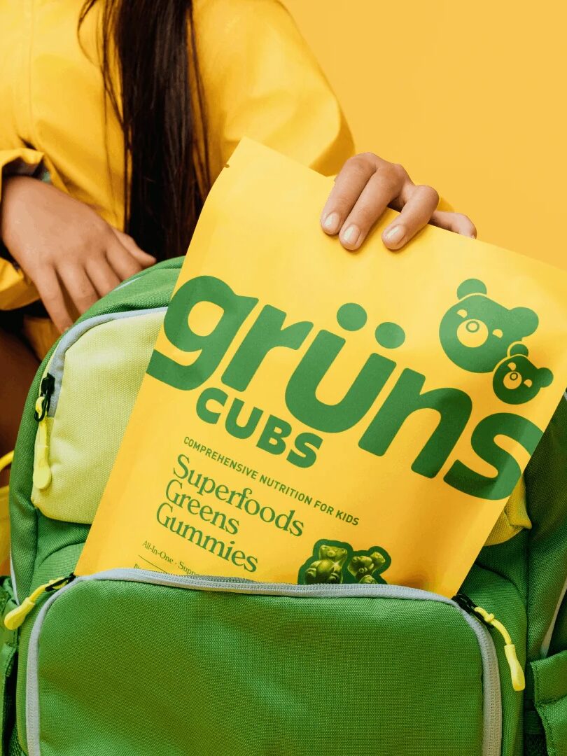 A person in a yellow jacket places a yellow bag of Grüns Cubs Superfoods Greens Gummies into a green backpack against a yellow background.