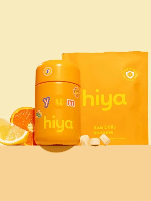 Orange container and packet labeled "hiya" with "yum" text, surrounded by a sliced orange, lemon, and chewable tablets, against a yellow background.