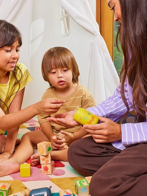 Two children and an adult sit on a play mat with wooden blocks. The adult offers vitamins from a yellow bottle to the children.
