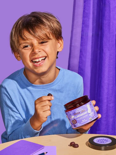 Smiling boy in a blue shirt holds a jar of blueberry-flavored products and a piece of candy at a table with a purple background.