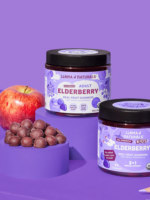 Two jars of elderberry gummies, an apple, grapes, a glass of ice water, and colored pencils are arranged on a purple surface with a purple background.