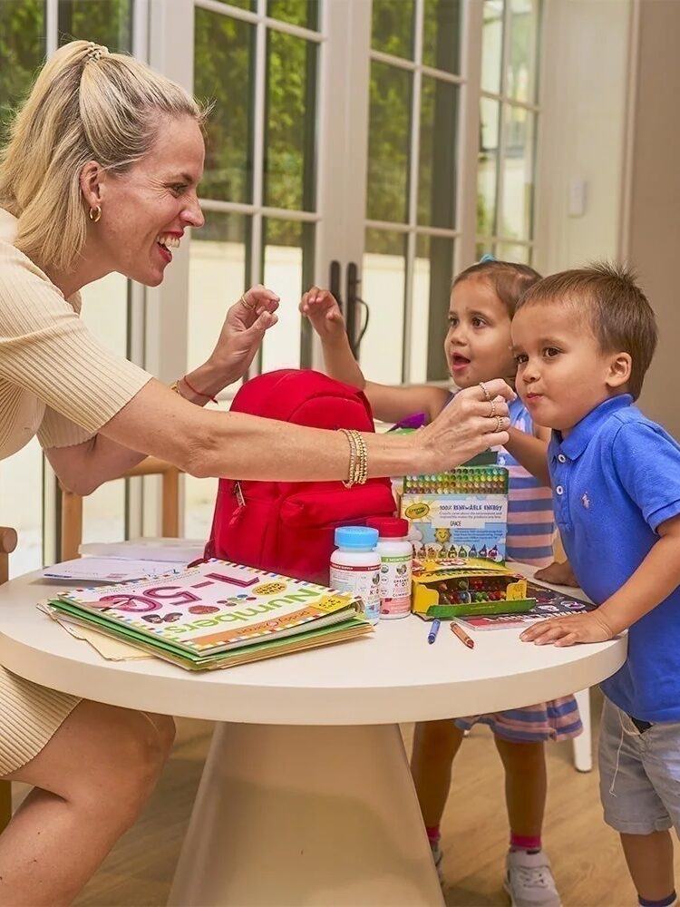 A woman interacts with two young children at a round table with books, a red bag, and a bottle in a brightly lit room.