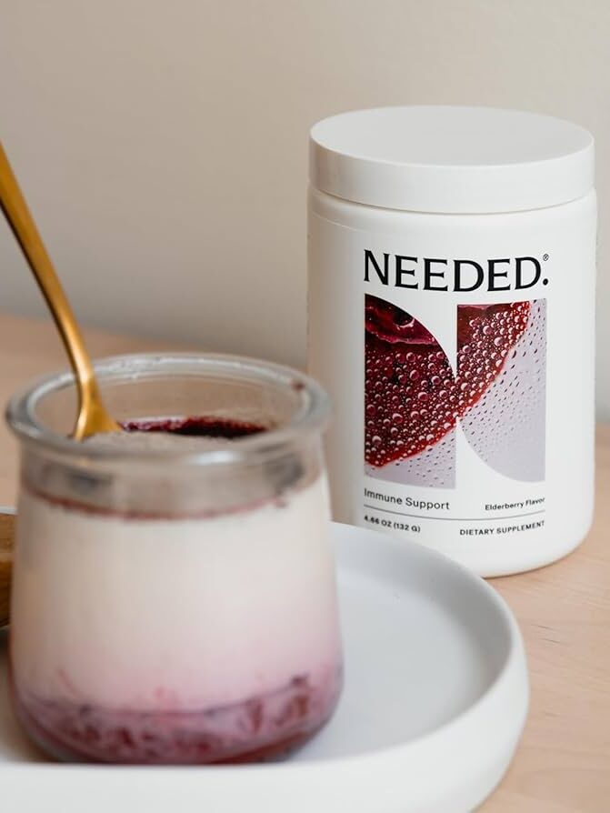 A jar of yogurt with berry sauce and a gold spoon sits beside a container labeled “NEEDED: Immune Support Elderberry Powder” on a white tray.
