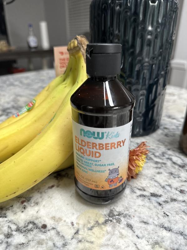 A bottle of NOW Kids Elderberry Liquid is placed on a marble countertop next to a bunch of bananas and a dark vase.