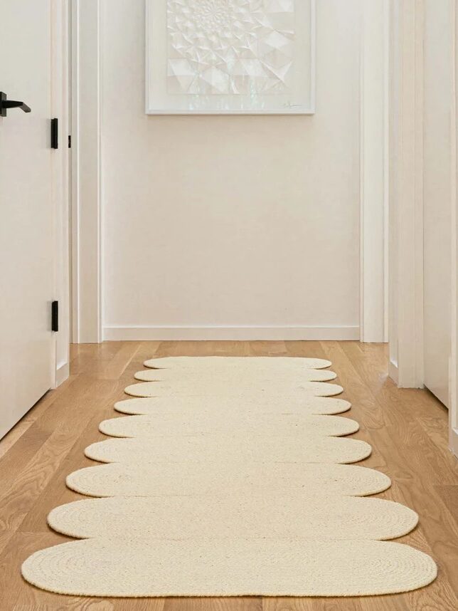 A beige scalloped-edge runner rug is laid on a wooden floor in a narrow hallway with white walls and doors.