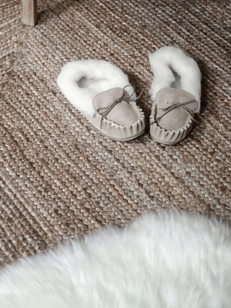 A pair of beige, fur-lined slippers rests on a textured woven rug next to the edge of a white furry mat.