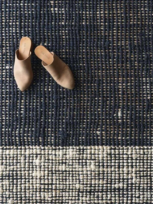 A pair of beige slip-on shoes placed on a textured woven rug with a dark blue and white color pattern.