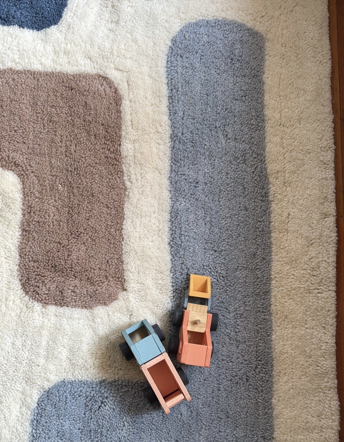 Two small wooden toy trucks, one blue and one orange, sit on a patterned rug with blue, beige, and brown shapes.