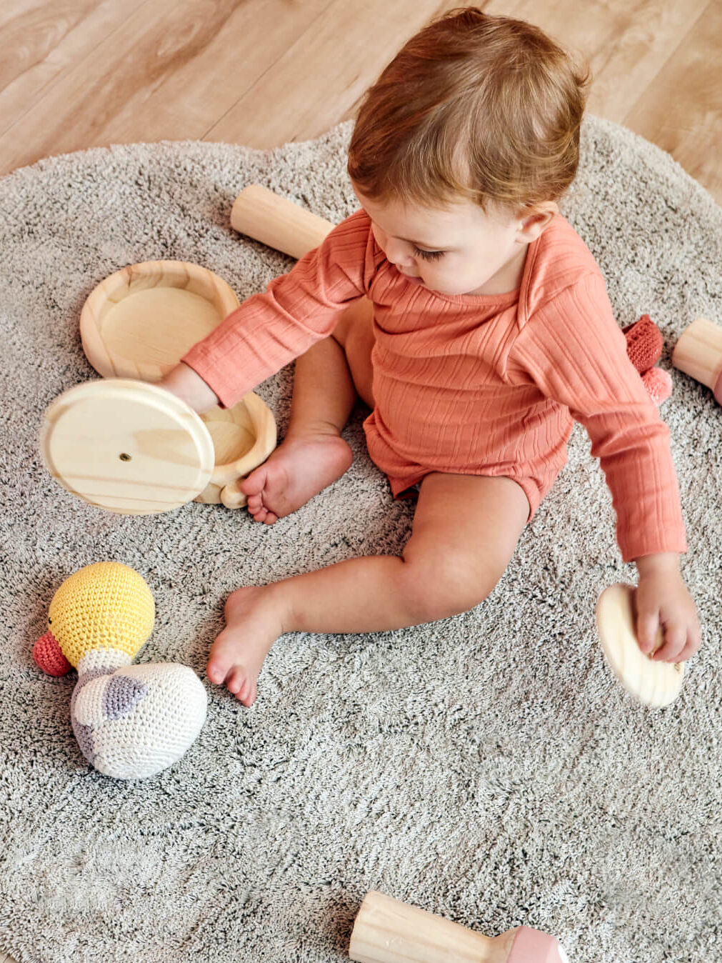 A toddler sits on a gray carpet playing with wooden toys and knitted balls on a light wood floor.