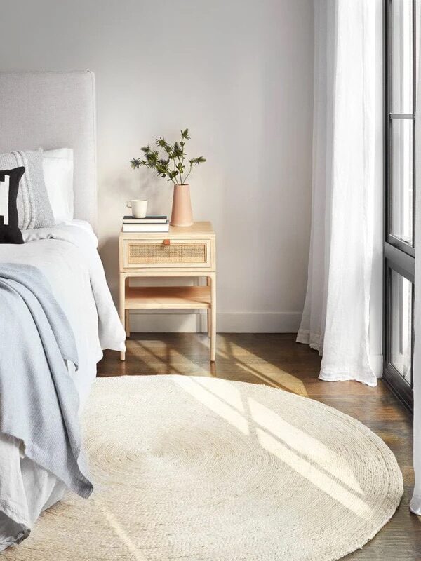A modern bedroom with a light gray bed, black and white pillow, woven basket, wooden nightstand with vase, and sunlight streaming through sheer curtains onto a round rug.