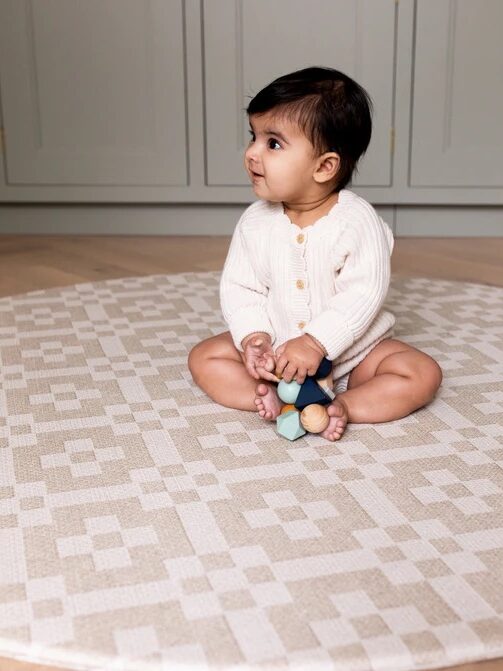 A baby in a white sweater sits on a patterned rug holding a small toy, looking to the side.
