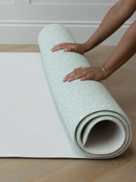 A person rolls out a light green patterned yoga mat on a wooden floor.