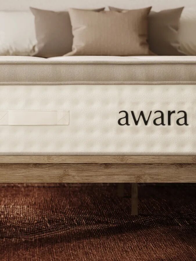 A bed with an Awara mattress on a wooden frame in a modern bedroom with a rug, pillows, and a nightstand holding a vase with flowers.