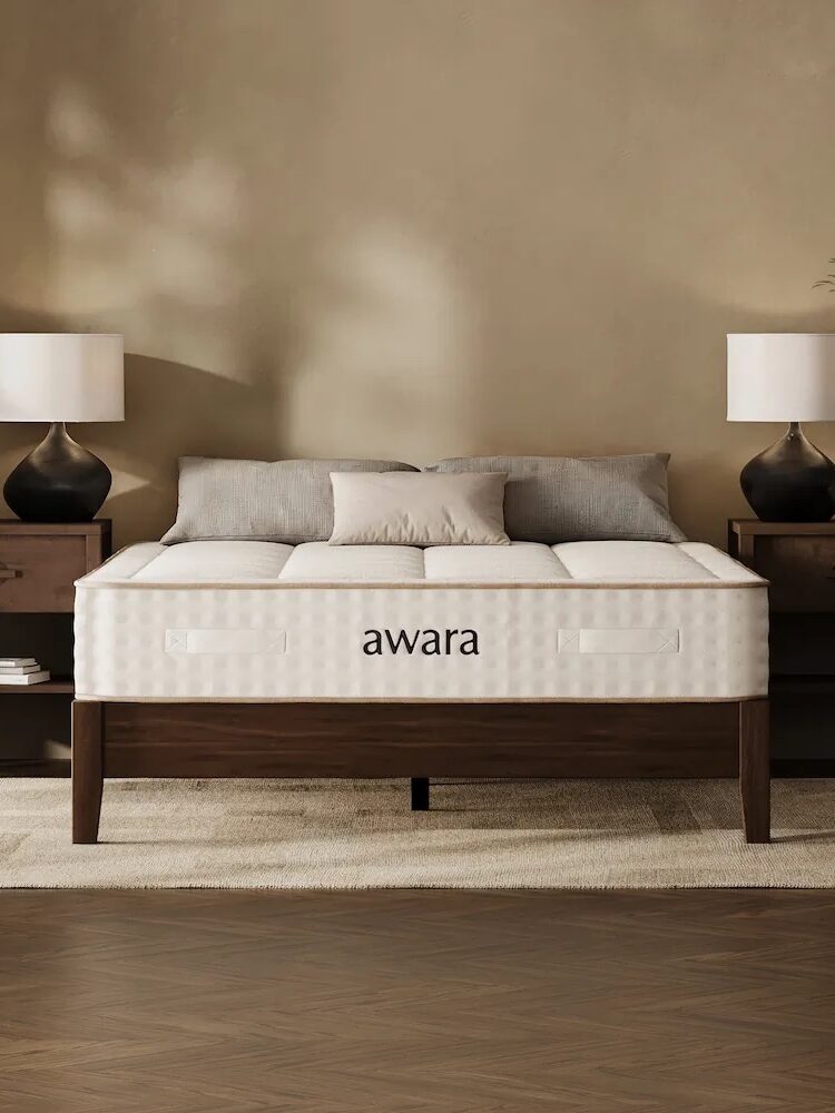 A neatly made bed with an "awara" mattress, flanked by two wooden nightstands with matching lamps, set against a beige wall with a rug underneath.