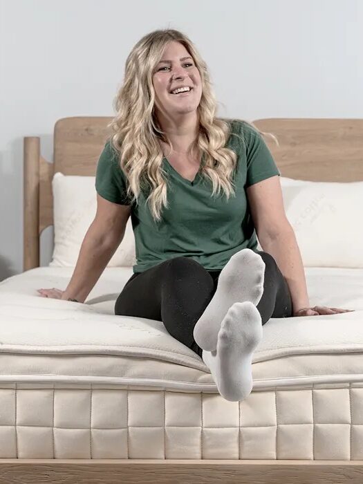 A woman with long blonde hair sits on a bed, smiling, with legs extended and feet pointed toward the camera.