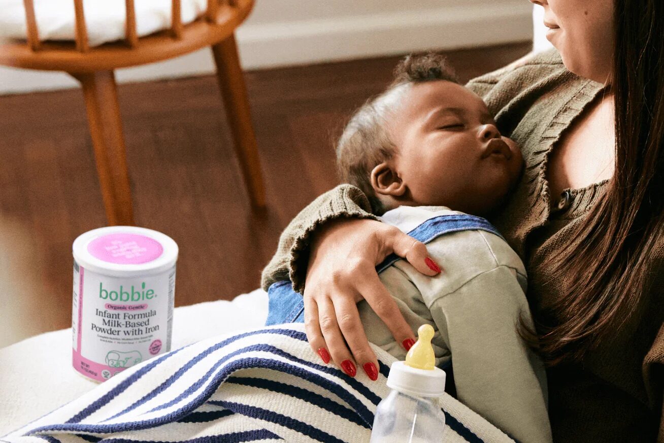 A woman holds a sleeping baby on her lap, with a baby bottle and a can of Bobbie infant formula nearby. Logos for purity and organic certifications are displayed at the bottom, highlighting the Labor Day Sale.