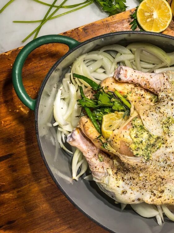 Whole seasoned chicken in a Dutch oven on a bed of sliced onions, herbs, and lemon, with a bowl of herb butter nearby—perfect for your next Labor Day sale gathering. Green lid set aside.
