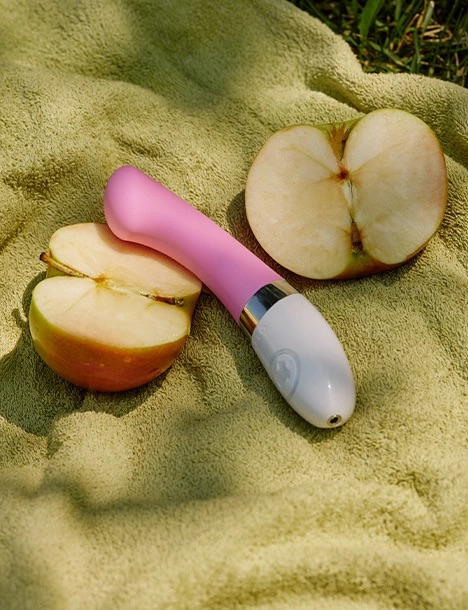 A pink and white vibrator placed on a towel outdoors next to two halves of a sliced apple.