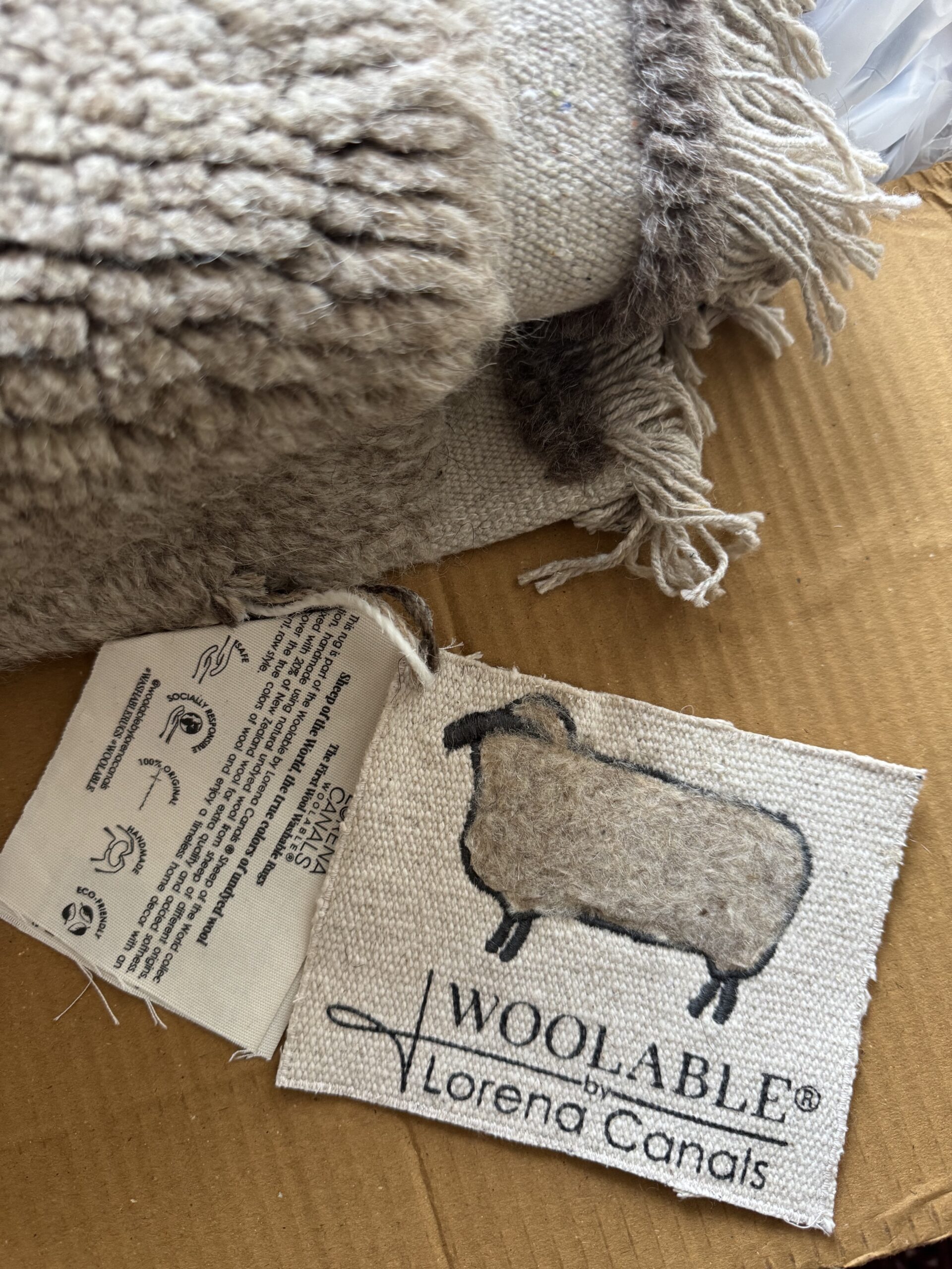 A close-up of a wool rug with fringed edges, a care label, and a fabric tag featuring a sheep illustration and the text "WOOLABLE by Lorena Canals.