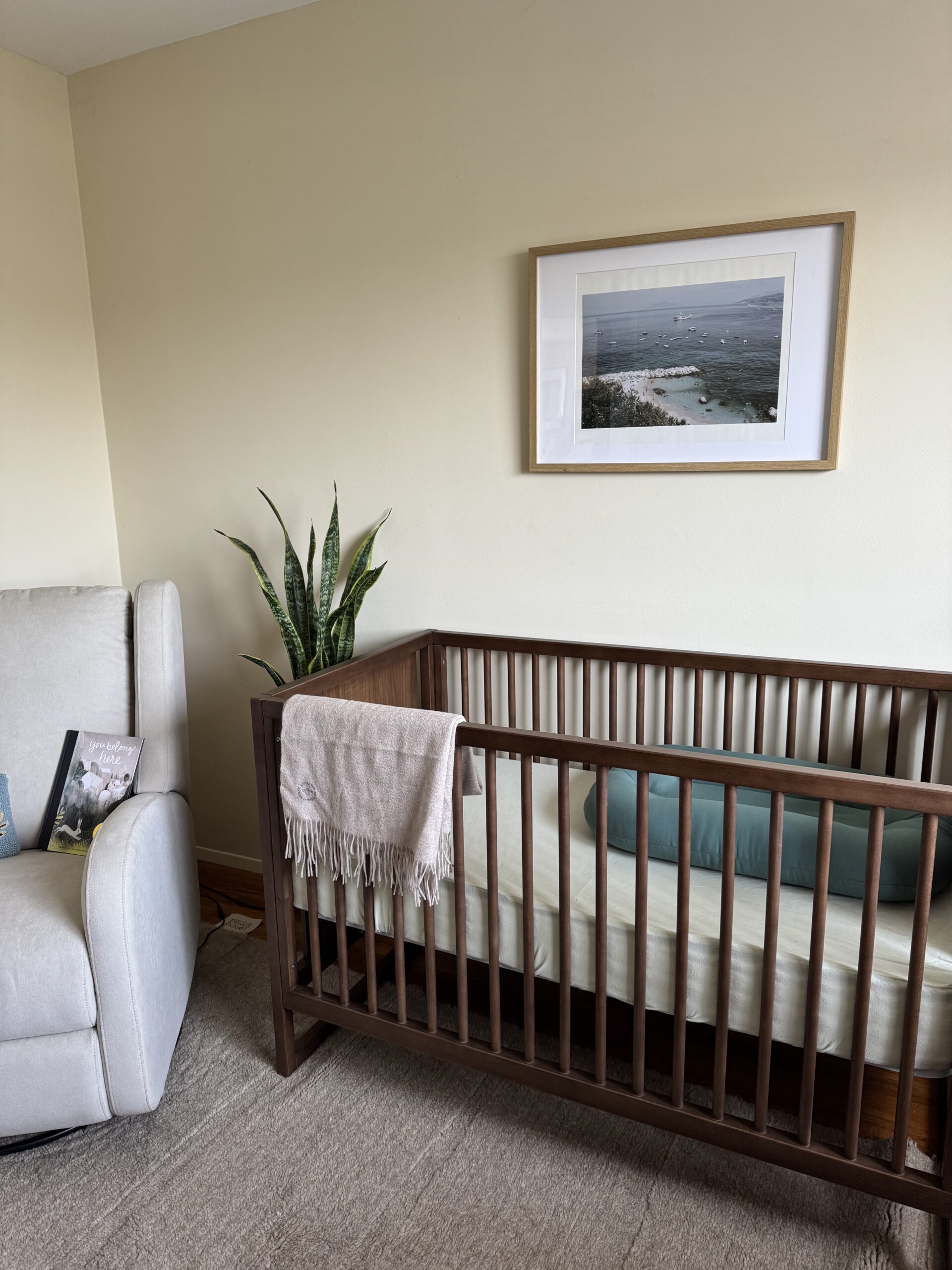 A wooden crib with a light blanket draped over the side sits next to a green plant and a gray armchair in a softly lit nursery. A framed photo hangs on the wall above.