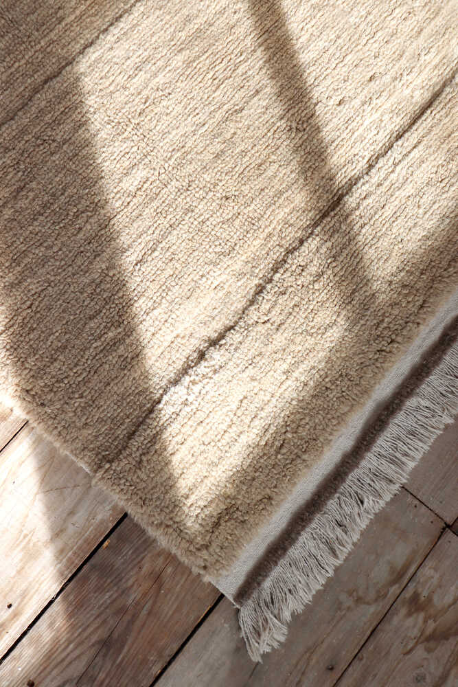 A beige textured rug with fringe on the edge lies on a wooden floor, with sunlight casting shadows across its surface.