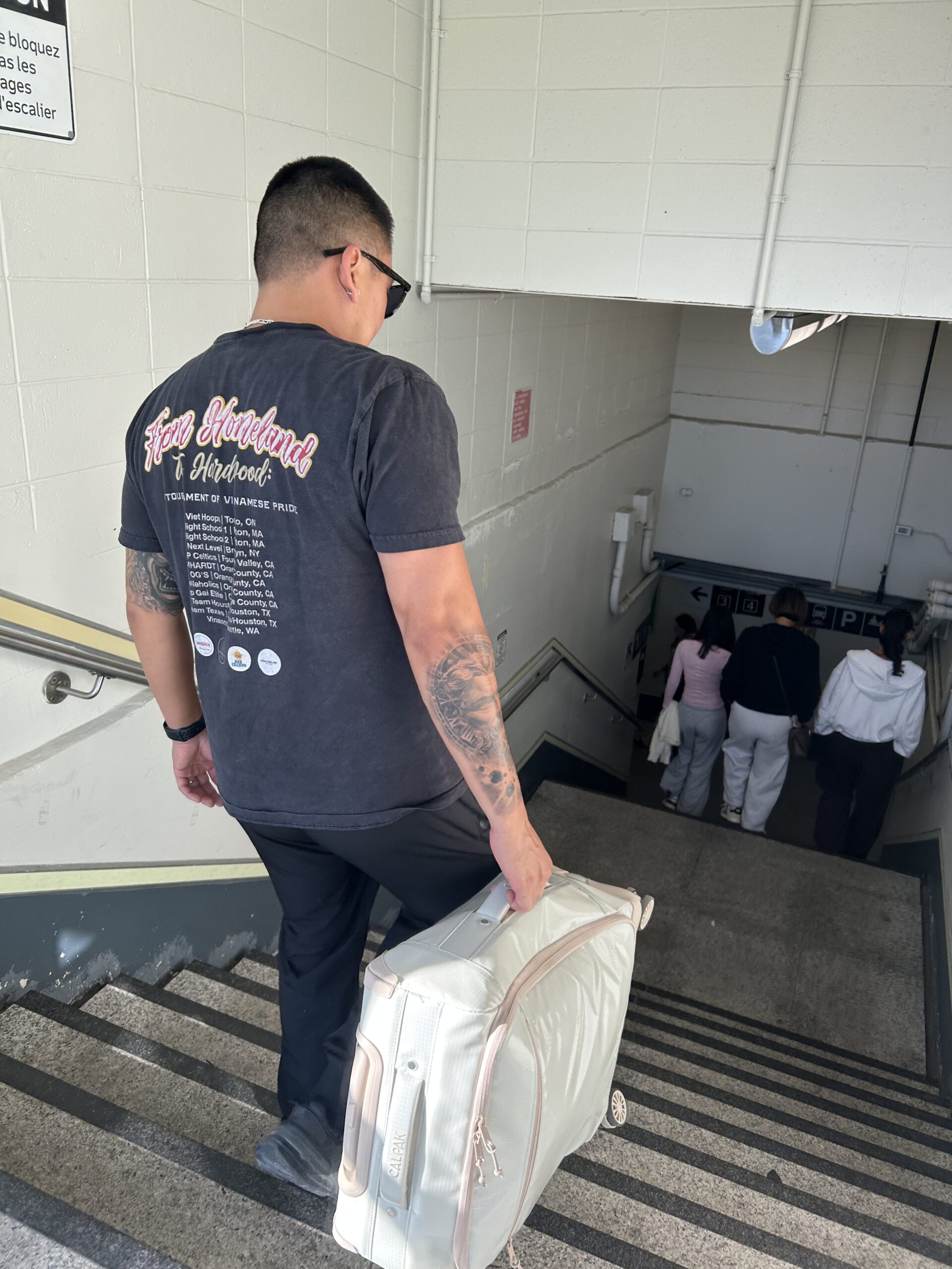 A man wearing a black T-shirt and sunglasses carries a large suitcase down a stairwell, following three people ahead of him.