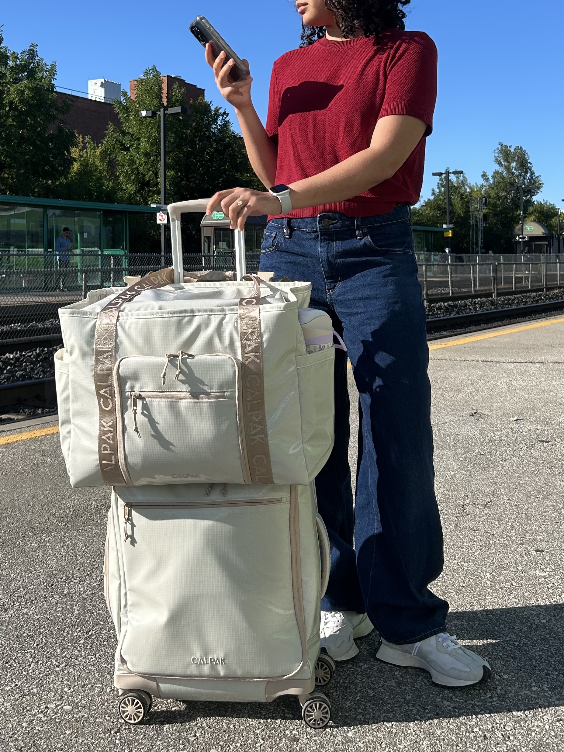 A person in a red shirt and blue jeans stands on a train platform, holding a phone and a large beige rolling suitcase with a matching bag stacked on top.