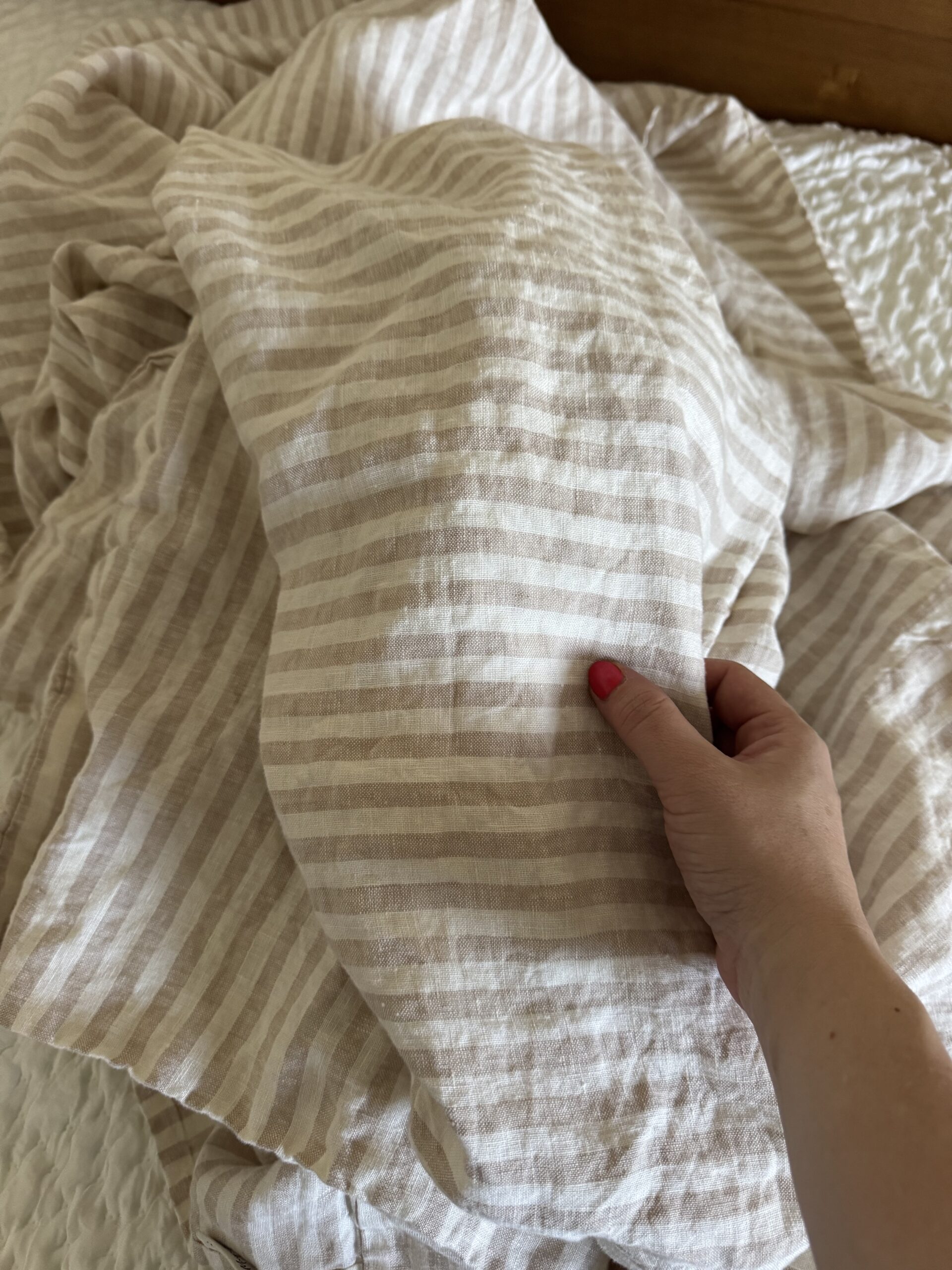 A hand with pink nail polish holds a piece of beige and white striped fabric draped over a bed with a white textured blanket.