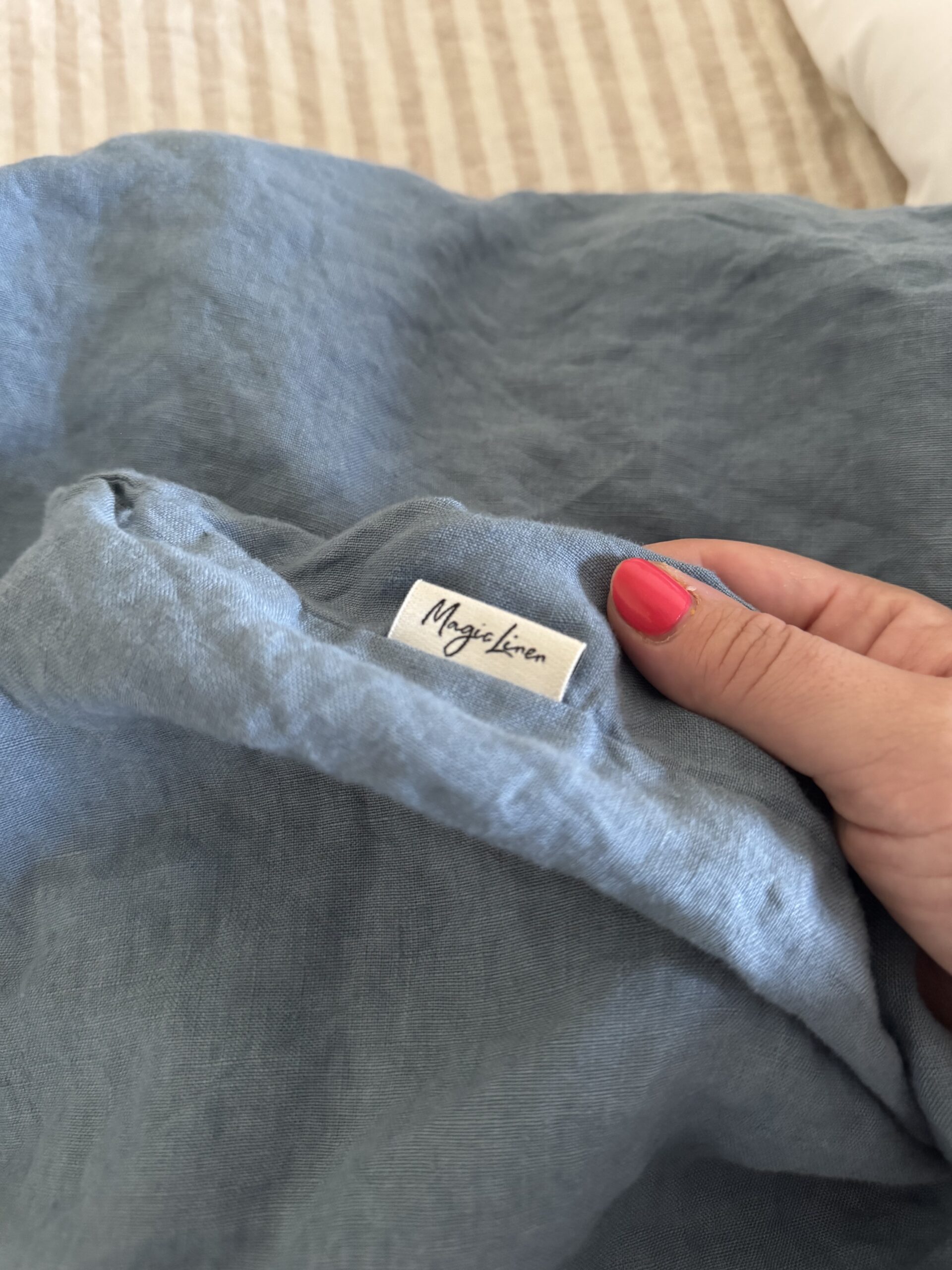 A hand with pink nail polish holds a piece of light blue fabric labeled “MagicLinen.” A beige and white striped fabric is in the background.