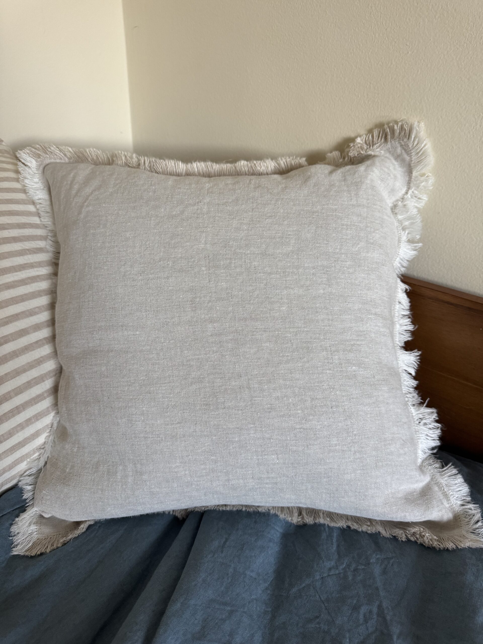 Square beige pillow with frayed edges on a bed with a blue sheet, next to a striped pillow and a wooden headboard against a cream wall.