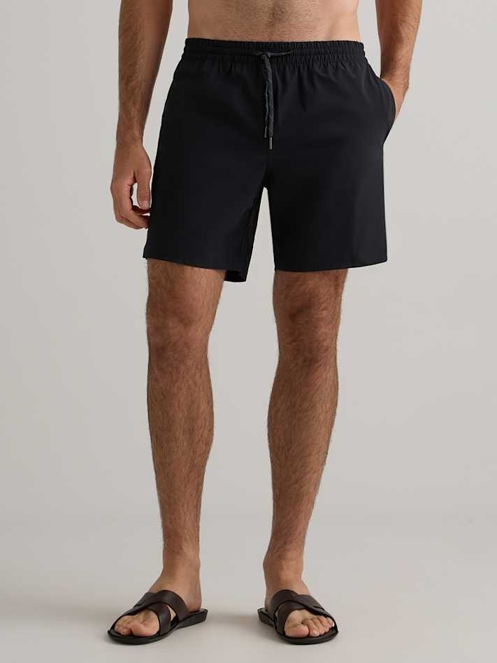 A man wearing black drawstring shorts and brown sandals stands against a plain light gray background.