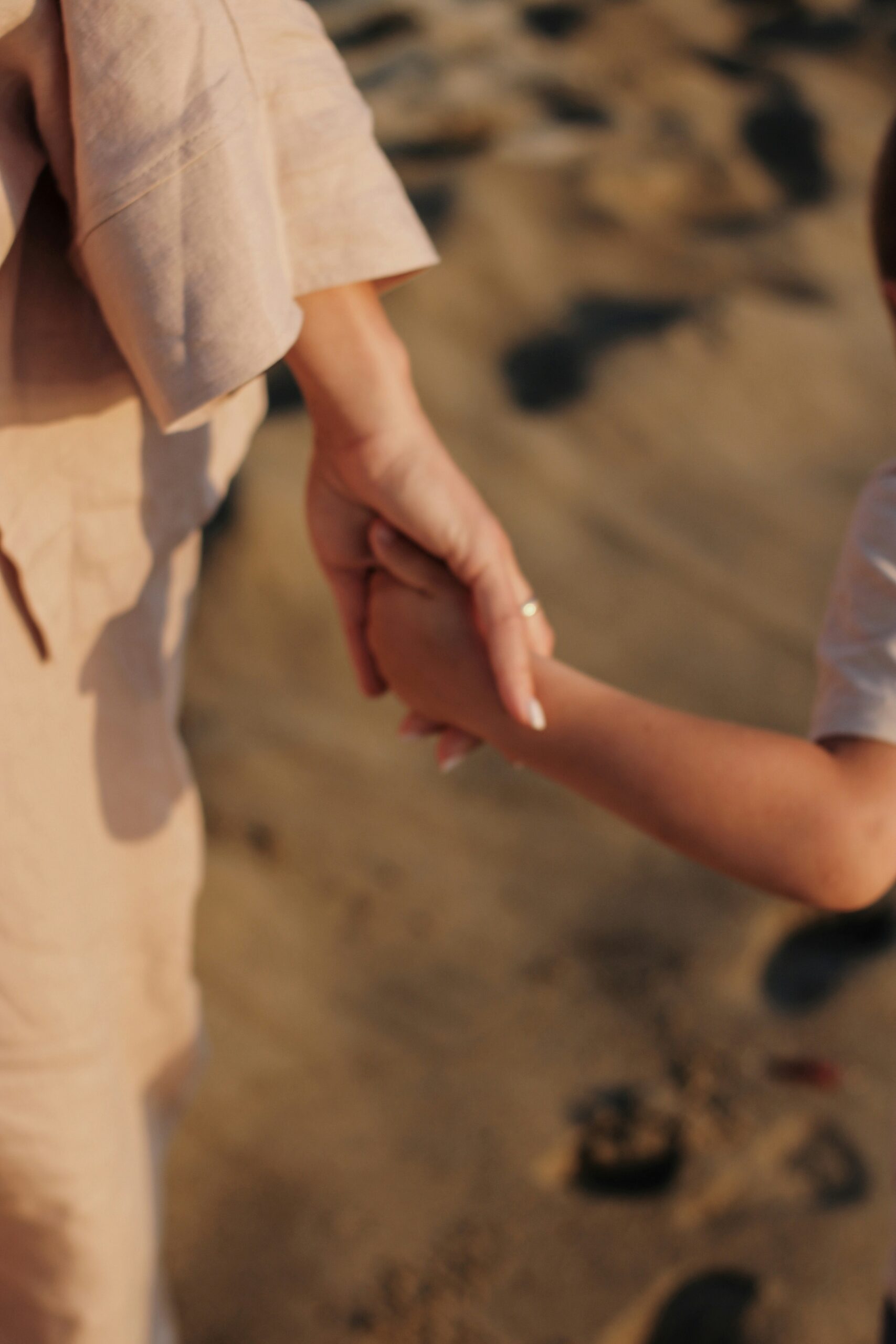 An adult and a child hold hands while standing on a sandy surface.