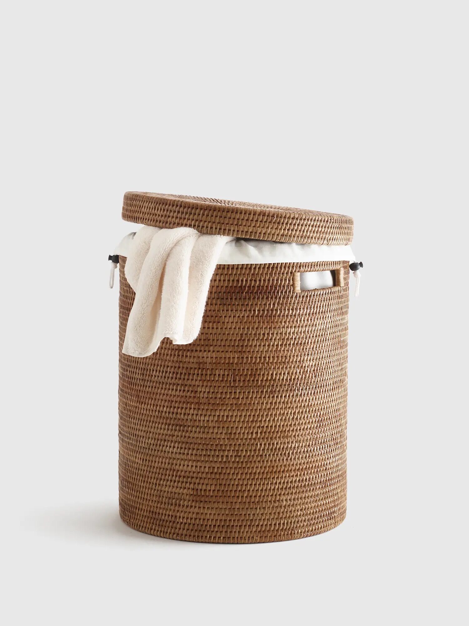 A round woven wicker laundry basket with a lid, slightly open, and a white towel draped over the edge against a plain background.