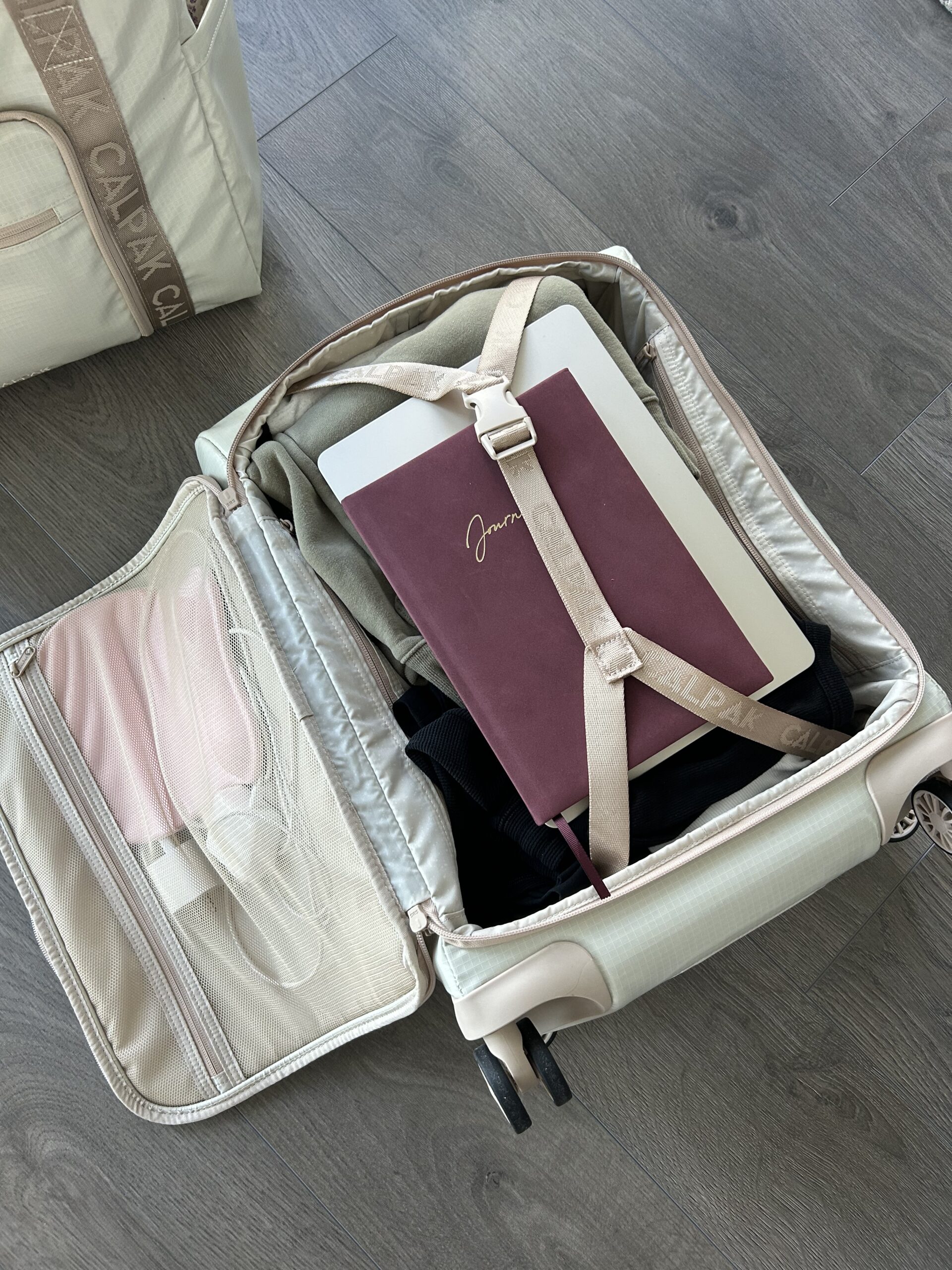 An open suitcase on a wooden floor contains neatly packed clothes, a closed laptop, and a maroon journal secured by straps.