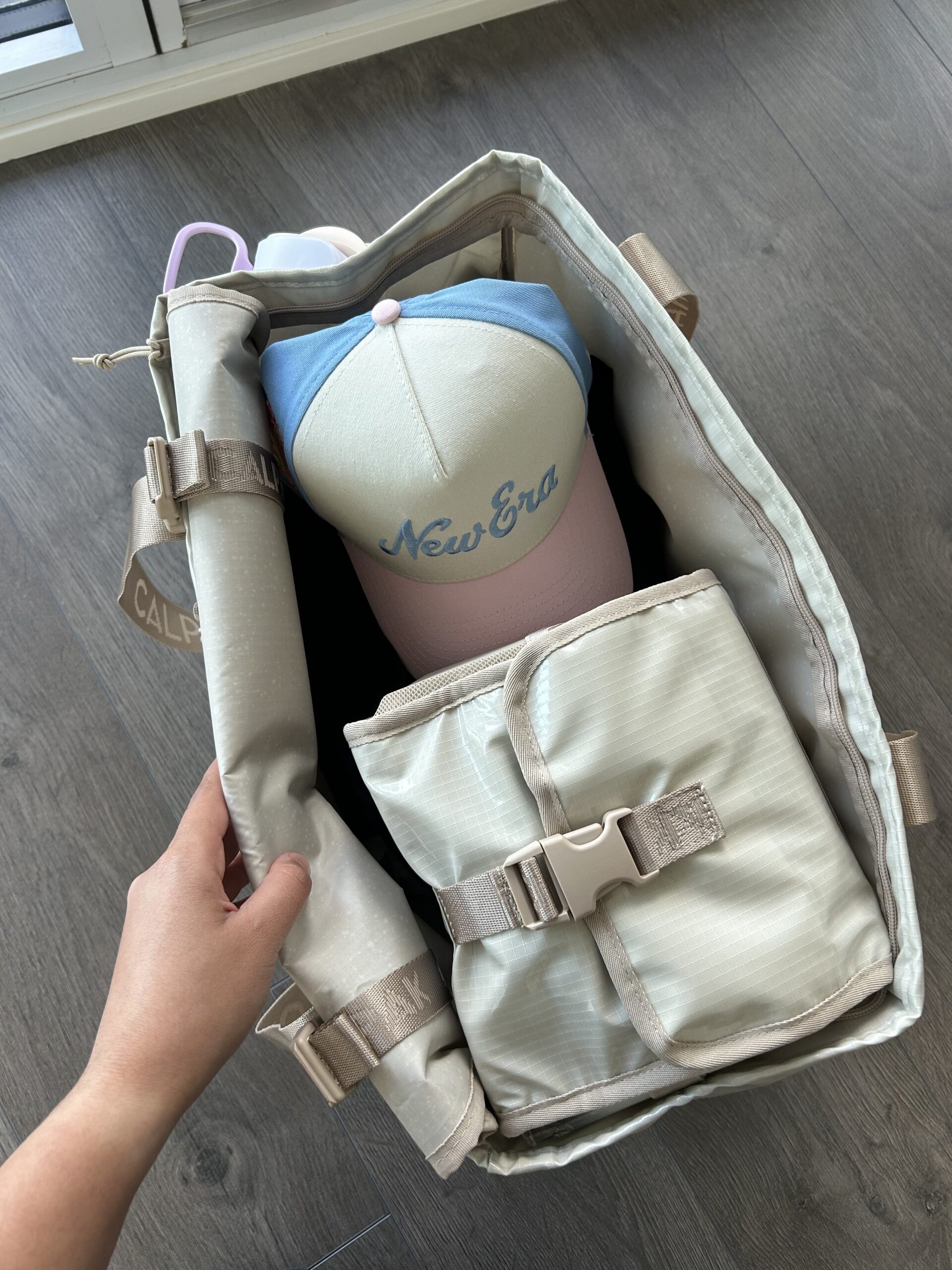 An open beige bag with a blue and white "New Era" cap and a folded, buckled changing pad inside, on a wooden floor.