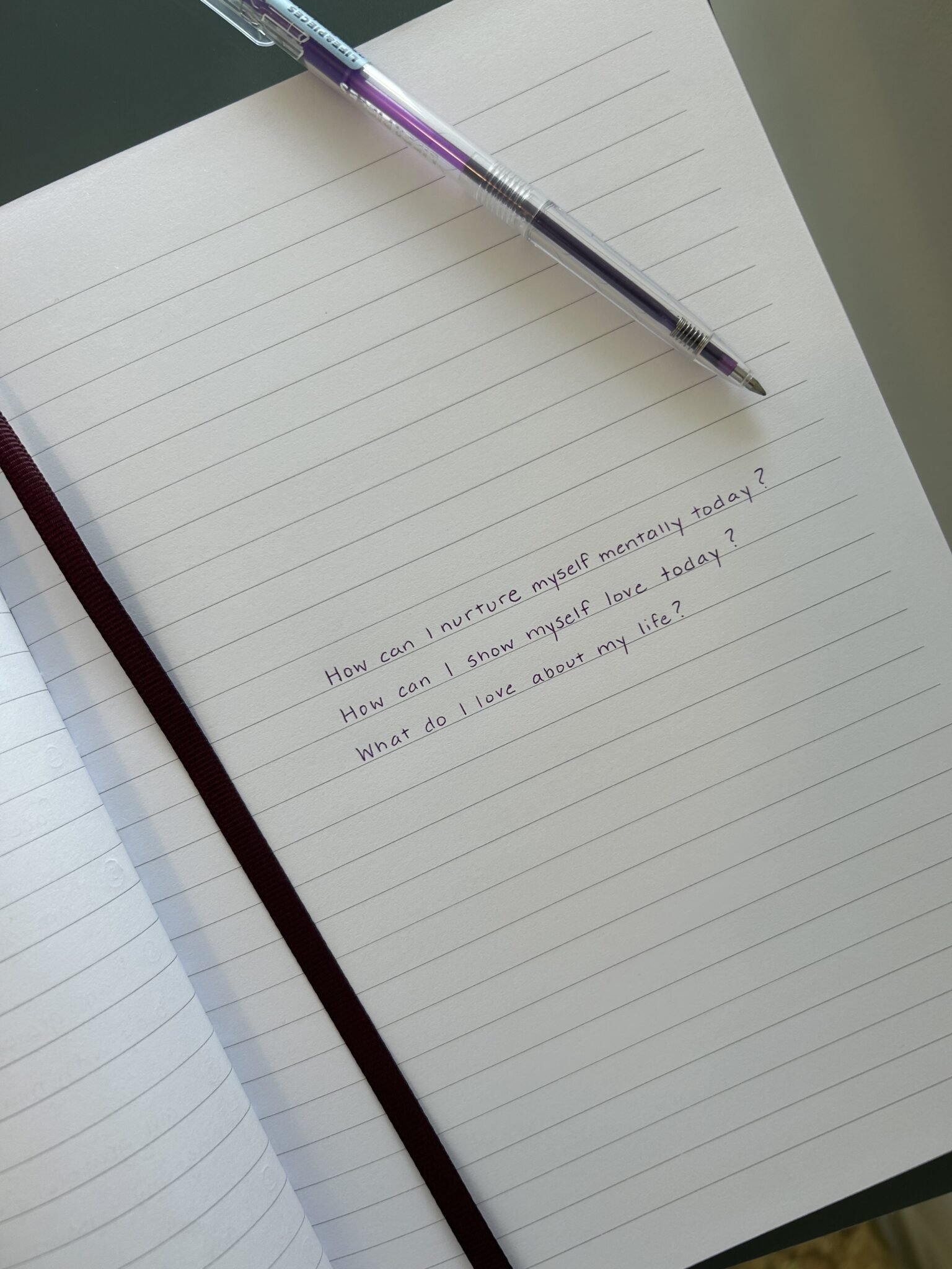 An open notebook with a maroon ribbon bookmark and a pen resting on it. The page has three handwritten self-reflection questions about self-care and self-love.