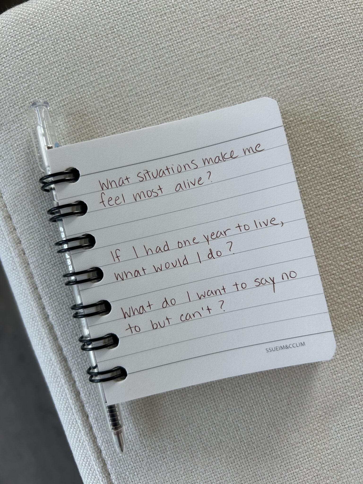 A spiral notebook with handwritten prompts: "What situations make me feel most alive?", "If I had one year to live, what would I do?", and "What do I want to say no to but can't?.