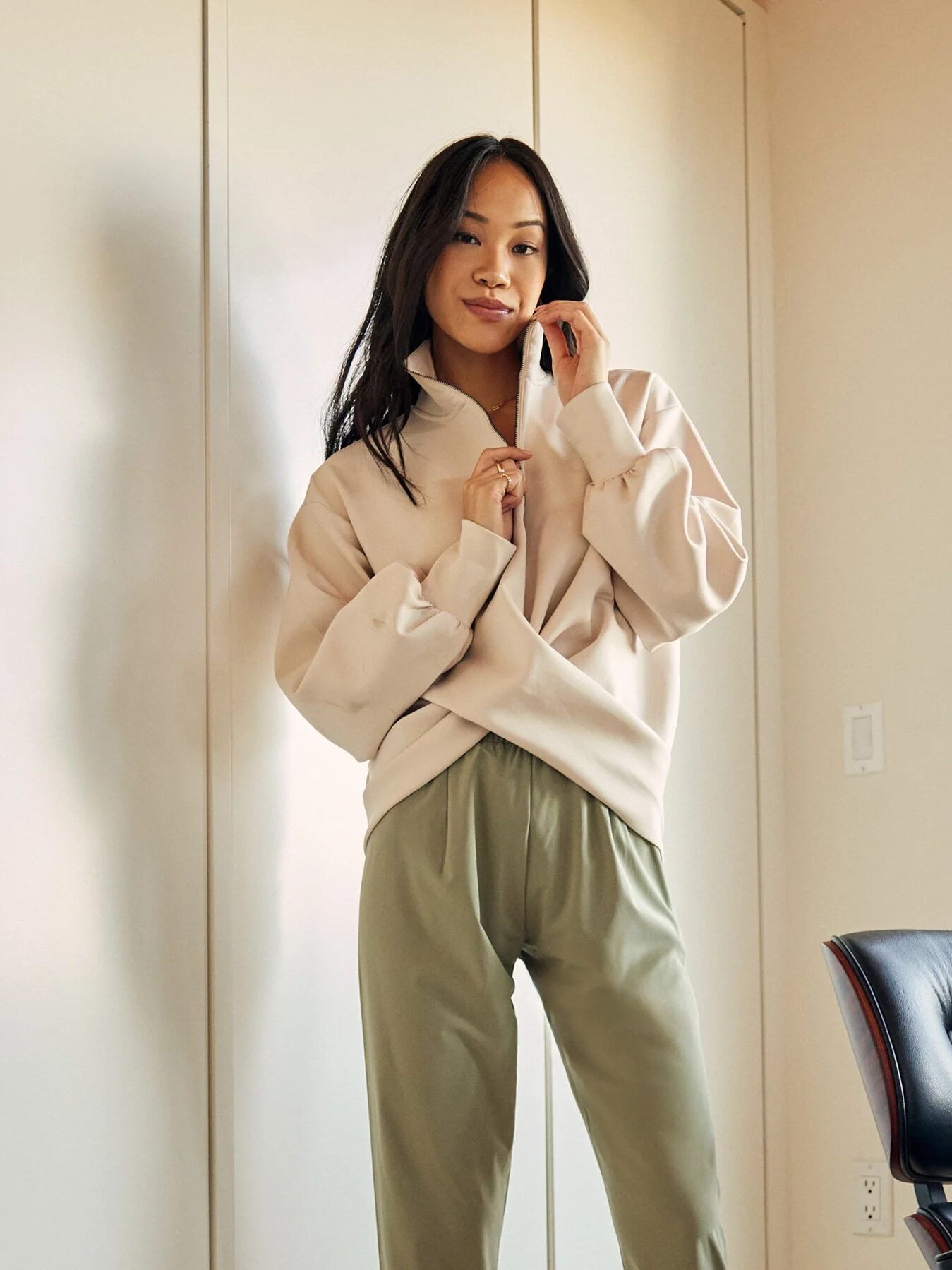 A person stands indoors wearing a beige pullover and olive green pants, with one hand touching their collar. A black chair is partially visible on the right.