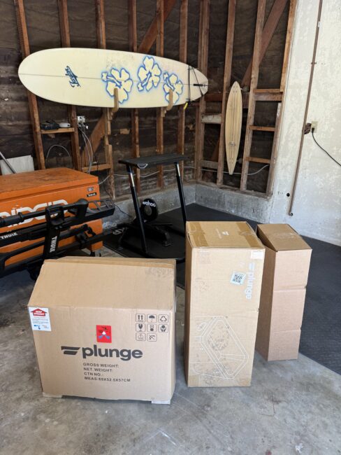 Four cardboard boxes, one labeled "Plunge," are on the floor of a garage with exposed wooden beams, a rowing machine, a Wahoo bike trainer, and two surfboards.